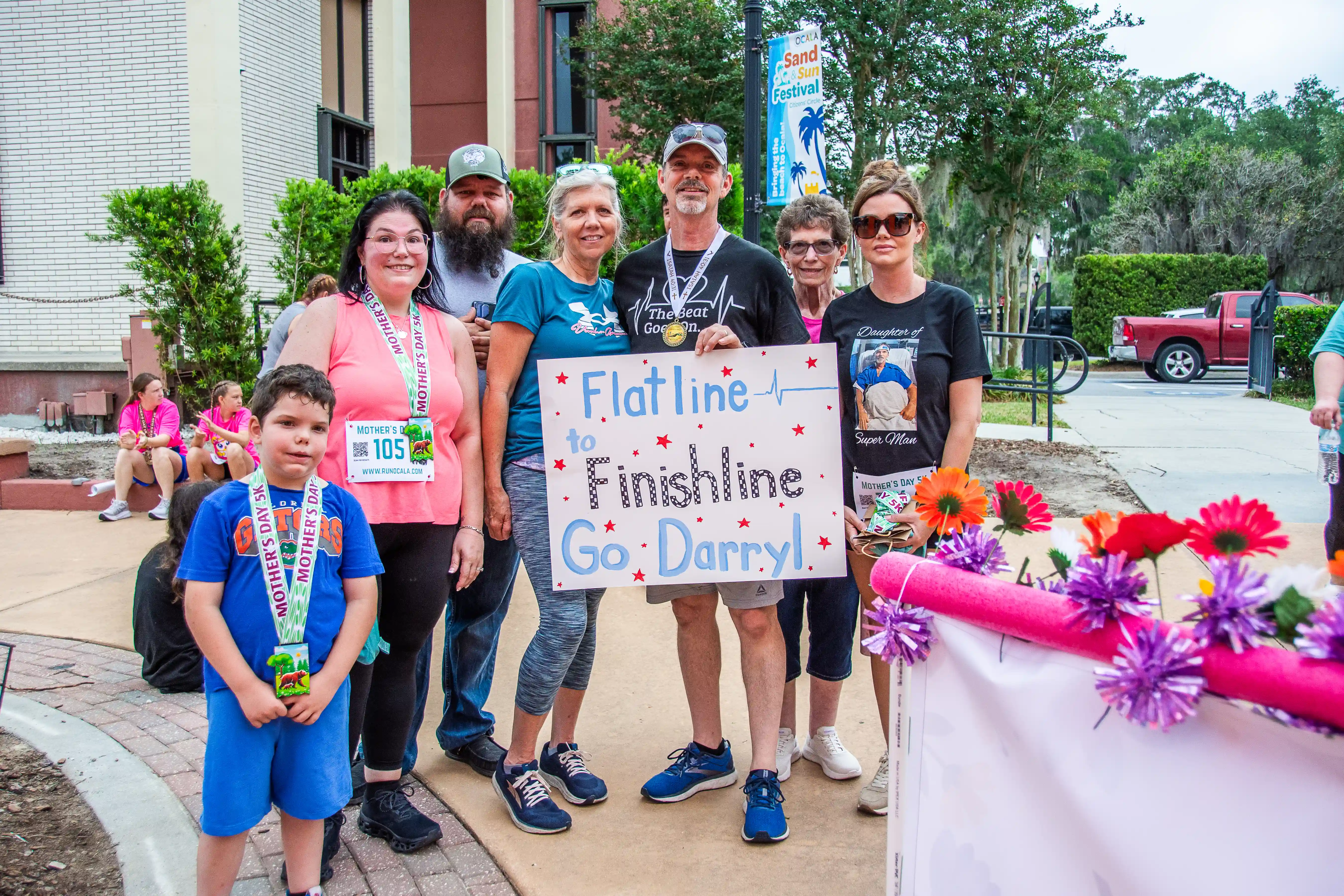 Darryl McGinnes wearing his Flatline to Finishline shirt and finisher medal at the 2026 Mother's Day 5K, surrounded by family and friends holding a hand-painted sign celebrating his return one year after suffering a heart attack at the same race